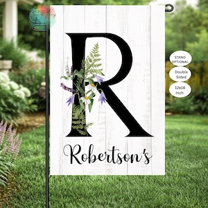 May include: A double-sided garden flag with a large black letter "R" and the name "Robertson's" in script. The letter is adorned with floral accents in purple, white, and green. The flag measures 12x18 inches and is displayed on a garden stand.