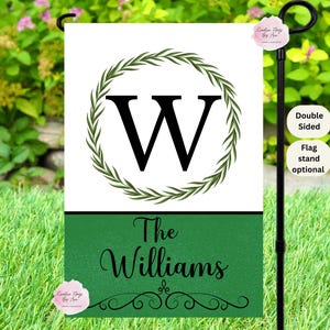 May include: A double-sided garden flag with a white background, featuring a black letter "W" encircled by a green wreath. The bottom section is green with the family name "The Williams" in black script.