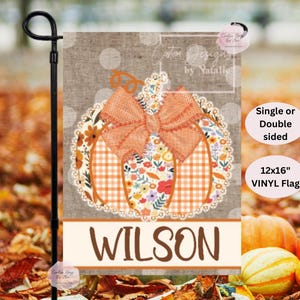 May include: A 12x16 inch vinyl garden flag featuring a pumpkin design with a bow and floral accents. The flag has the name "WILSON" at the bottom. The background is a fall-themed setting with pumpkins and leaves.
