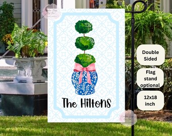 May include: A blue and white ceramic planter with a pink bow and three green topiary trees. The planter has a floral design and the text "The Hilltons" is printed below the planter. The flag is 12x18 inches and is double-sided.