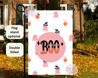 May include: A double-sided Halloween garden flag featuring a pink and orange design. The flag has a white background with pink ghosts, cupcakes, and the word "BOO" in a retro font. The flag is displayed on a black flag stand.