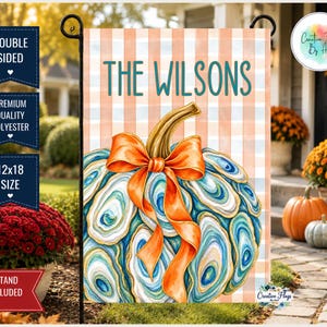 May include: A decorative garden flag featuring a pumpkin made of blue and white oyster shells with an orange ribbon bow. The background has a light orange and white checkered pattern. The text THE WILSONS is printed in teal at the top of the flag.