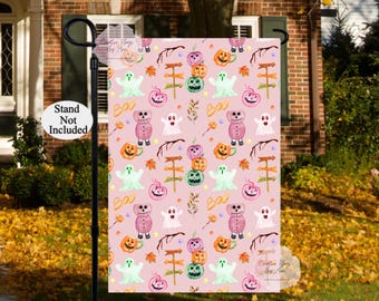 May include: A Halloween garden flag with a repeating pattern of ghosts, pumpkins, bears, and the word "Boo" on a light pink background. The flag is displayed outdoors, with a "Stand Not Included" sign visible.