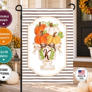 May include: A rectangular garden flag featuring a brown and white horizontal striped background. The center displays a white decorative frame containing an illustration of orange, white, and yellow pumpkins in a pedestal bowl with a brown bow and the letter K.