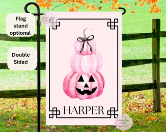 May include: A double-sided pink garden flag featuring a stack of pink pumpkins with a black bow and a carved jack-o'-lantern face. The flag has a black border and the name "HARPER" in black text. A flag stand is optional.