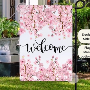 May include: A white welcome sign with pink cherry blossoms. The sign is double-sided and has a flag stand.