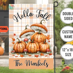 May include: A rectangular garden flag featuring a light orange and white plaid background. The text reads Hello Fall and The Markels. The design includes four orange pumpkins with smiling faces, several mushrooms, and scattered autumn leaves.