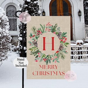 May include: A beige Christmas garden flag with a wreath design. The wreath features pine needles, red berries, and pine cones around a red circle with the letter "H". The words "MERRY CHRISTMAS" are printed in red below the wreath.