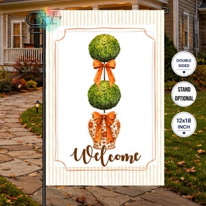 May include: A rectangular garden flag featuring a white background with a light tan striped border. The design displays two stacked green topiary balls in a patterned orange and white urn, accented with orange bows and the word Welcome in brown.