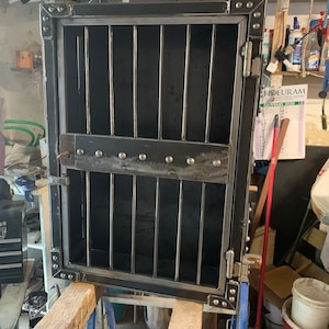 May include: A black metal cabinet with a door that has a metal grate. The cabinet has a hinge on the right side and a lock on the left side. The cabinet is sitting on a wooden table.