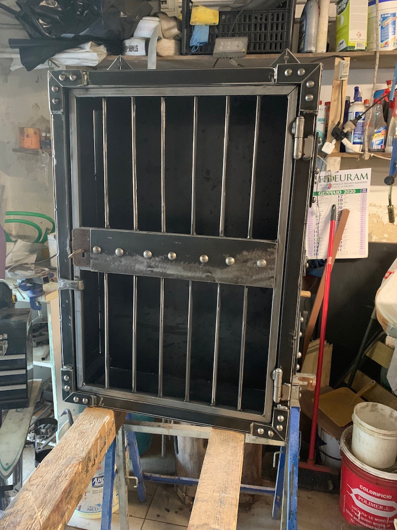 May include: A black metal cabinet with a door that has a metal grate. The cabinet has a hinge on the right side and a lock on the left side. The cabinet is sitting on a wooden table.