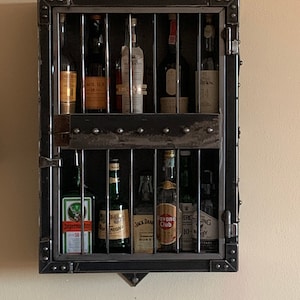 May include: A black metal wall mounted liquor cabinet with a caged door. The cabinet has two shelves and is filled with various bottles of liquor. The bottles include J&auml;germeister, Negroni, Jack Daniels, Havana Club, and more.