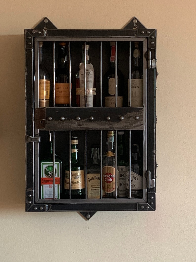 May include: A black metal wall mounted liquor cabinet with a caged door. The cabinet has two shelves and is filled with various bottles of liquor. The bottles include J&auml;germeister, Negroni, Jack Daniels, Havana Club, and more.