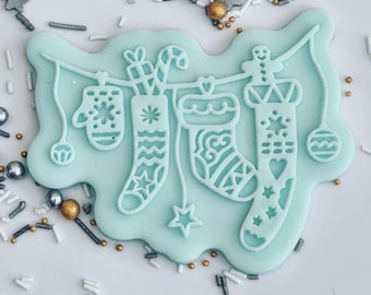 May include: A light blue cookie cutter in the shape of a garland with Christmas stockings, a mitten, and ornaments. The cookie cutter has intricate cut-out designs and is surrounded by silver and gold sprinkles.