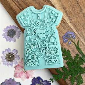 May include: A light blue cookie cutter shaped like a baby onesie with various designs including stars, hearts, a rainbow, a dog, a duck, a car, and the text "A New Baby To Love".