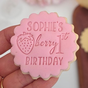 May include: A pink frosted cookie with a scalloped edge. The cookie is stamped with the words "Sophie's berry 1st birthday" and a strawberry illustration. The cookie is held in a hand.