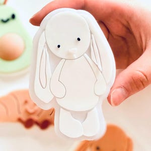 Peut inclure: Un emporte-pièce blanc en forme de lapin avec des yeux en points noirs et de longues oreilles est tenu dans une main. D'autres emporte-pièces en forme d'avocat, de croissant et d'un autre lapin sont visibles en arrière-plan. Les emporte-pièces sont probablement pour la pâtisserie.