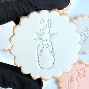 May include: A hand in a black glove holds a white iced cookie with a scalloped edge. The cookie has a light blue outline of a rabbit. Other cookies with rabbit and duck designs are visible.