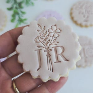 May include: A light pink, scalloped-edge biscuit with a floral design and the letters "J R" is held in a hand. Other biscuits with similar designs are in the background. The biscuits appear to be stamped with a floral design.