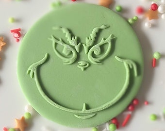Op de afbeelding: Een lichtgroene, cirkelvormige fondant met een reliëf Grinch-gezicht. De fondant is omgeven door kleurrijke hagelslag, waaronder rode, witte, groene en stervormige hagelslag. De afbeelding is een close-up.