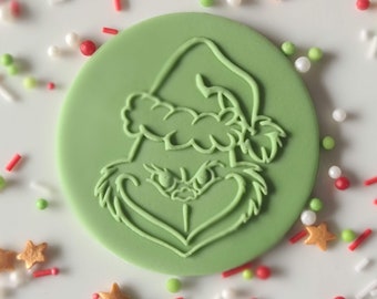 Op de afbeelding: Een ronde, lichtgroene koekjessteker met het gezicht van de Grinch met een kerstmuts. De steker is omringd door kleurrijke sprinkles, waaronder rode, groene, witte en stervormige sprinkles. Perfect voor kerstgebak.