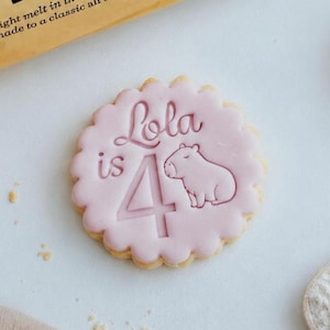 May include: A round, scalloped-edge biscuit with light pink icing, embossed with the text 'Lola is 4' and a small capybara illustration. Beside it lies a gold-coloured M&S Food packet for all butter biscuits, featuring black text and nutritional data.