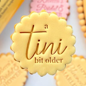 May include: Close-up of several round, scalloped-edge cookies. One cookie has the text "a tini bit older" engraved on it. Other cookies have text, including "Rebecca's Bridal & Shower" and "Happy Blessed Marlini".