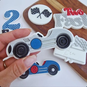 Puede incluir: Un conjunto de decoraciones comestibles para un pastel de cumpleaños, que incluye un coche de carreras plateado, un coche de carreras azul, una bandera a cuadros, un número dos y las palabras "Two Fast".