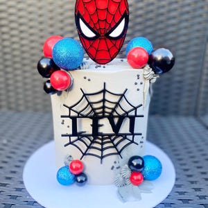 Puede incluir: Un pastel blanco decorado con globos rojos, azules y negros, una telara&ntilde;a negra y una placa de nombre negra 'LEVI'. Un topper de m&aacute;scara de Spiderman rojo y negro se coloca en la parte superior del pastel.