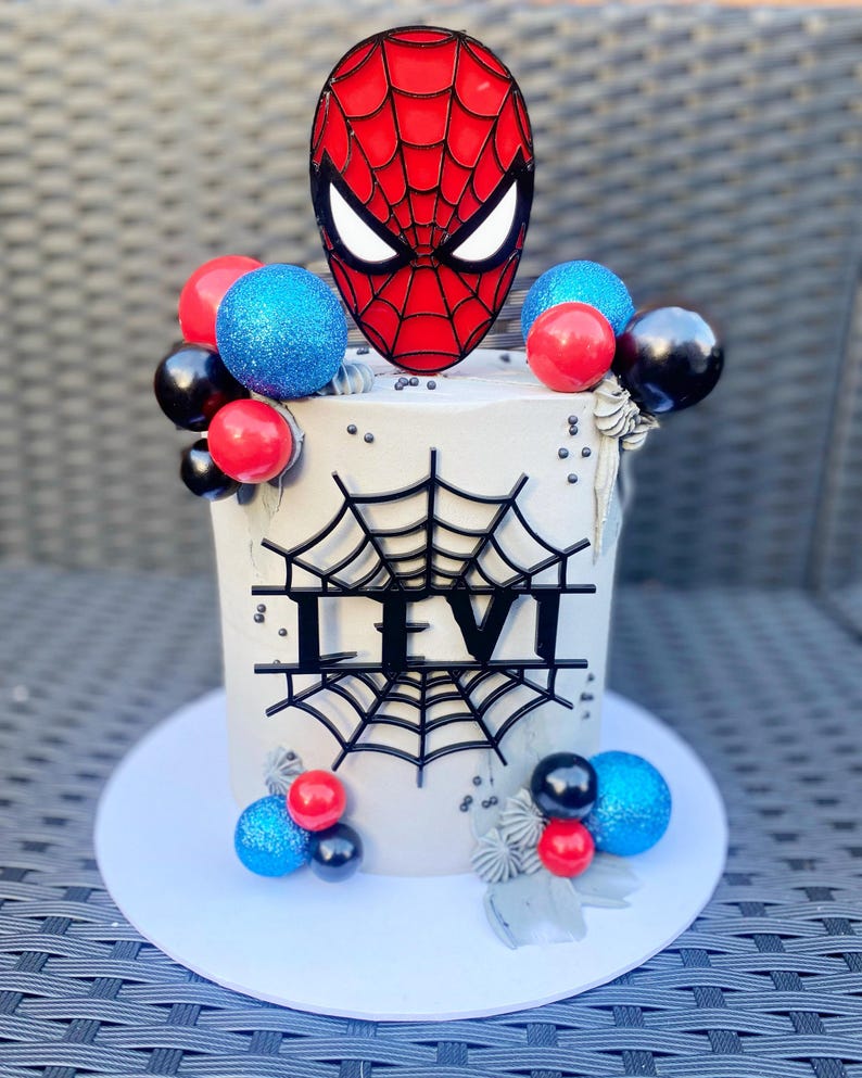 Puede incluir: Un pastel blanco decorado con globos rojos, azules y negros, una telara&ntilde;a negra y una placa de nombre negra 'LEVI'. Un topper de m&aacute;scara de Spiderman rojo y negro se coloca en la parte superior del pastel.