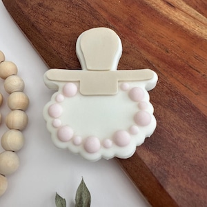 May include: A white pacifier cookie decorated with a pink pearl necklace. The cookie is on a wooden surface.