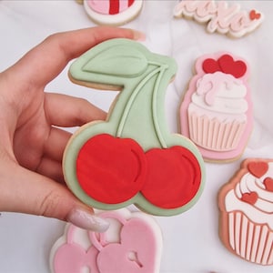 Puede incluir: Una galleta de azúcar verde y roja con forma de cereza con una hoja. La galleta está decorada con glaseado rojo y verde.
