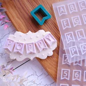 May include: A white cookie with a pink icing banner that reads "POPPY" and a white polka dot background. The cookie is on a wooden surface next to a clear plastic sheet with letters and numbers in a banner shape. There is also a green plastic cookie cutter in the shape of a banner.