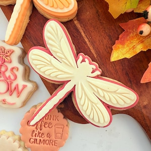 May include: A decorated cookie in the shape of a dragonfly with a cream-colored body and red outline. Other cookies include an orange slice, a leaf, and one with the text "Drink coffee like a Gilmore".