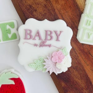 Może przedstawiać: Białe ciasteczko ozdobione różowym lukrem i napisem "BABY in bloom". Ciasteczko jest ozdobione zielonymi i różowymi kwiatami.