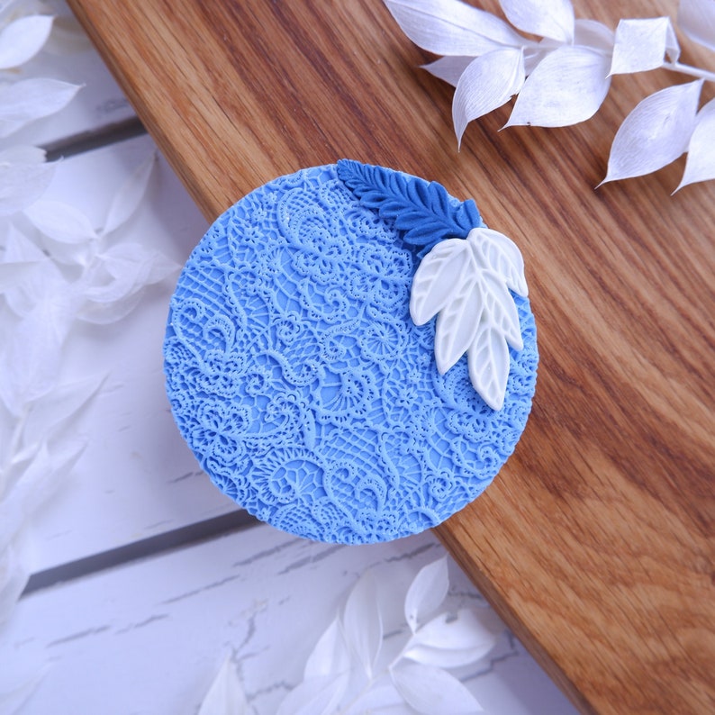 Op de afbeelding: Een ronde blauwe taarttopper met een wit en blauw kantpatroon en witte bladeren.