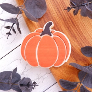 May include: A close-up of a pumpkin-shaped cookie cutter on a wooden surface with eucalyptus leaves. The cookie cutter is made of white ceramic and has a detailed design.