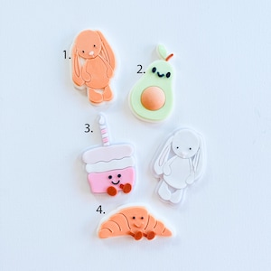 Peut inclure: Une collection de breloques colorées de style dessin animé. Les breloques comprennent un lapin orange, un avocat vert, un cupcake rose avec une bougie, un lapin blanc et un croissant orange. Chaque breloque a une bordure blanche.