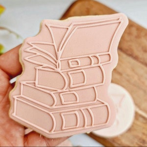 Könnte beinhalten: Ein rosa Zuckerkeks, der mit einem Stapel Bücher verziert ist. Der Keks ist in Form eines Bücherstapels geformt, wobei das oberste Buch geöffnet ist. Der Keks ist mit weißem Zuckerguss verziert.
