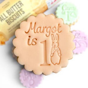 May include: A light peach-colored cookie with a scalloped edge, embossed with the words "Margot is 1" and a bunny illustration. Other cookies in pastel colors are in the background, along with a package of biscuits.