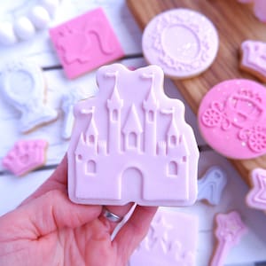 May include: A pink sugar cookie decorated with a white outline of a castle. The cookie is being held by a hand.