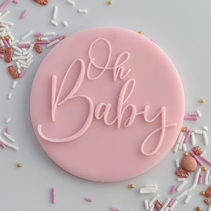 May include: A pale pink, circular cake topper with the words "Oh Baby" in a cursive script. The topper is surrounded by sprinkles in various colours, including white, pink, and gold. Ideal for a baby shower celebration.