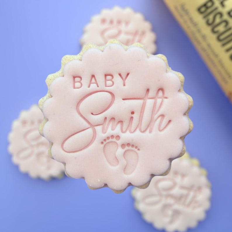 May include: Pink frosted sugar cookies with the name "Smith" and the word "Baby" imprinted on them, along with baby footprints. The cookies have a scalloped edge and are arranged on a blue surface.