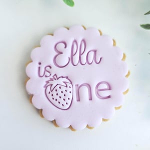 May include: A round biscuit with a scalloped edge. The biscuit has a light purple icing with the words "Ella is one" and a strawberry illustration. The biscuit is a light brown colour.