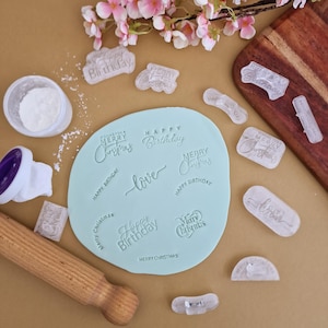 Può includere: Pasta di zucchero azzurra con testo in rilievo che recita "Happy Birthday", "Merry Christmas" e "love". Timbri trasparenti e un mattarello sono disposti attorno alla pasta di zucchero. Fiori rosa e un tagliere in legno sullo sfondo.
