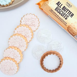 May include: A collection of baby shower cookies with the name "Taylor" in pink icing, along with a package of "All Butter Biscuits". The cookies have a scalloped edge and bear and elephant designs.