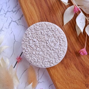 May include: A white round silicone stamp with a detailed floral design. The design features a variety of flowers and leaves in a circular pattern.