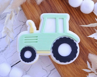 May include: A green tractor cookie cutter with black wheels and a yellow exhaust pipe. The cookie cutter is on a wooden surface.