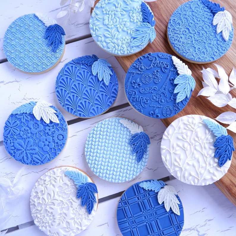 Op de afbeelding: Negen ronde blauwe en witte fondantkoekjes met verschillende reli&euml;fmotieven, waaronder bloem-, geometrische en bladpatronen. De koekjes zijn gerangschikt op een wit oppervlak met een houten snijplank op de achtergrond.