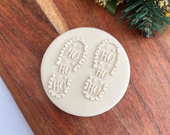 Op de afbeelding: Een ronde, crèmekleurige koekjesstempel met twee voetafdrukken met de tekst "HO HO HO". De voetafdrukken zijn versierd met sneeuwvlokken en sterren. De stempel ligt op een houten oppervlak met kerstversiering op de achtergrond.